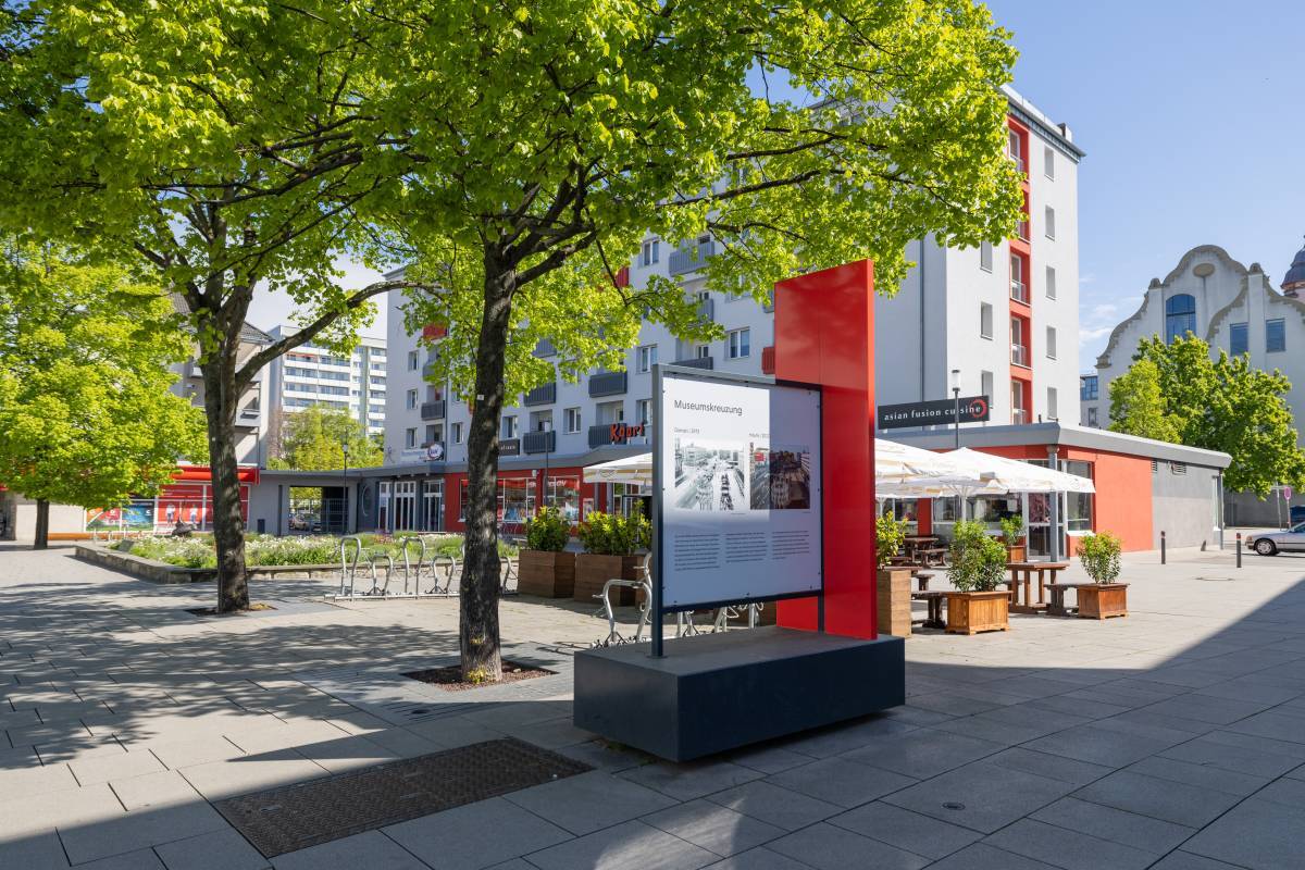 Großes rechteckiges Gebäude mit roten Akzenten an der Fassade sowie begrünter Ess- und Sitzbereich mit rotem Infoschild im Vordergrund.