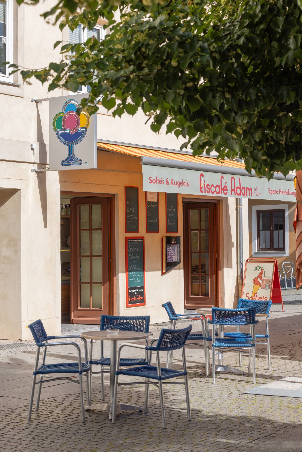 Außenbereich des Eiscafé Adam Dessau mit blauen Sitzgelegenheiten und mehreren Angebotstafeln sowie ein buntes Eislogo an der Wand.