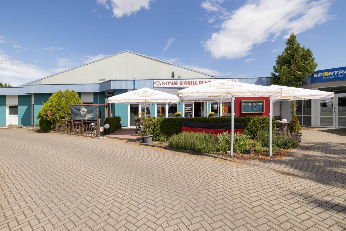 Begrünter Außenbereich des Restaurant Gusto Dessau mit offenen weißen Sonnenschirmen und blauer Fassade