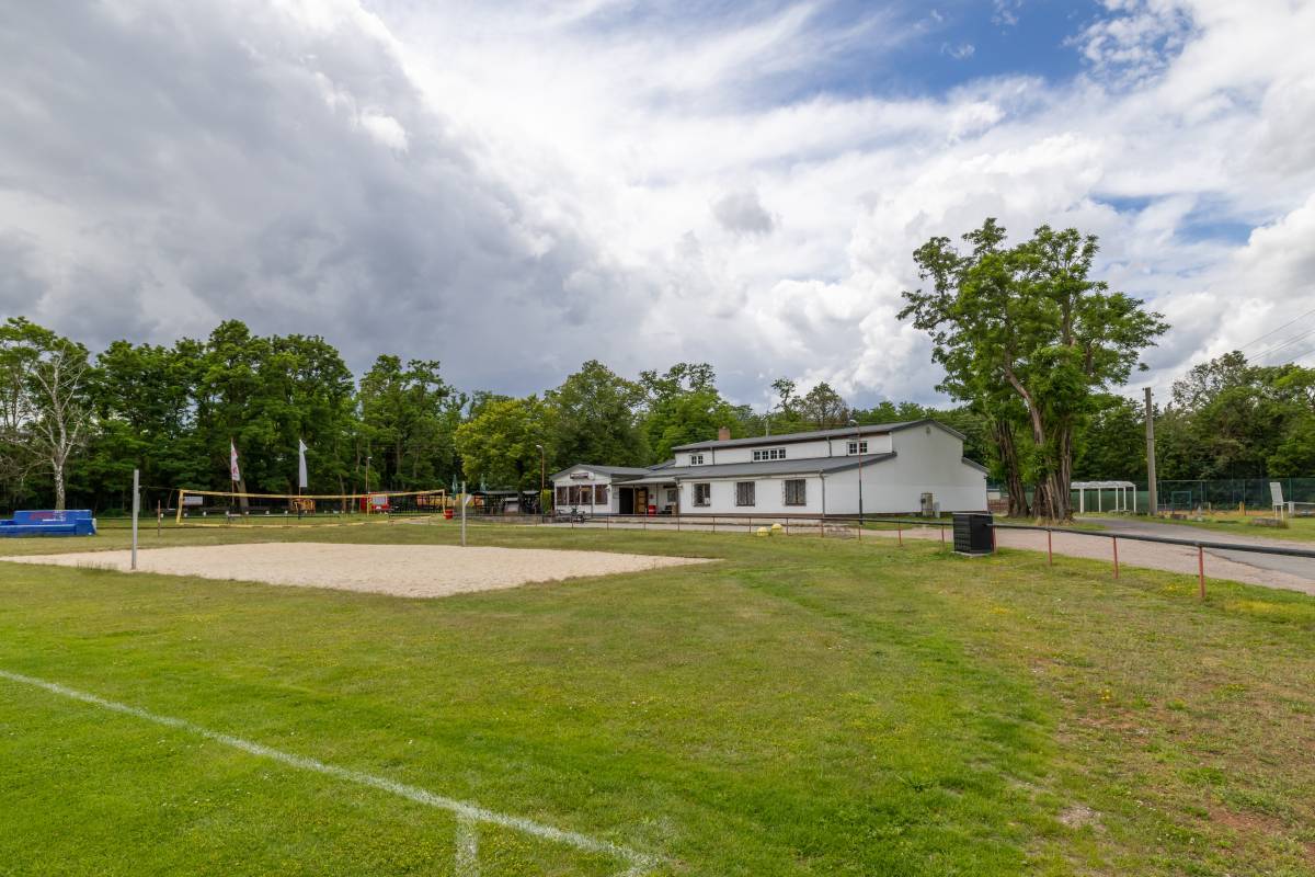 Begrünte Sicht auf die Einfahrt und das weiße Gebäude des Restaurant Lokheim mit Grünfläche und Volleyballplatz im Vordergrund