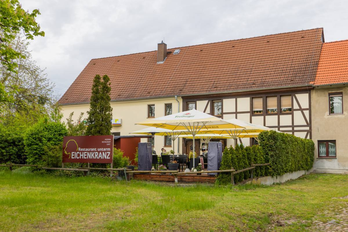 Begrünter Außensitz- und Essbereich des Restaurant unterm Eichenkranz mit gelb-weißen Sonnenschirmen und dem Restaurantschild davor.