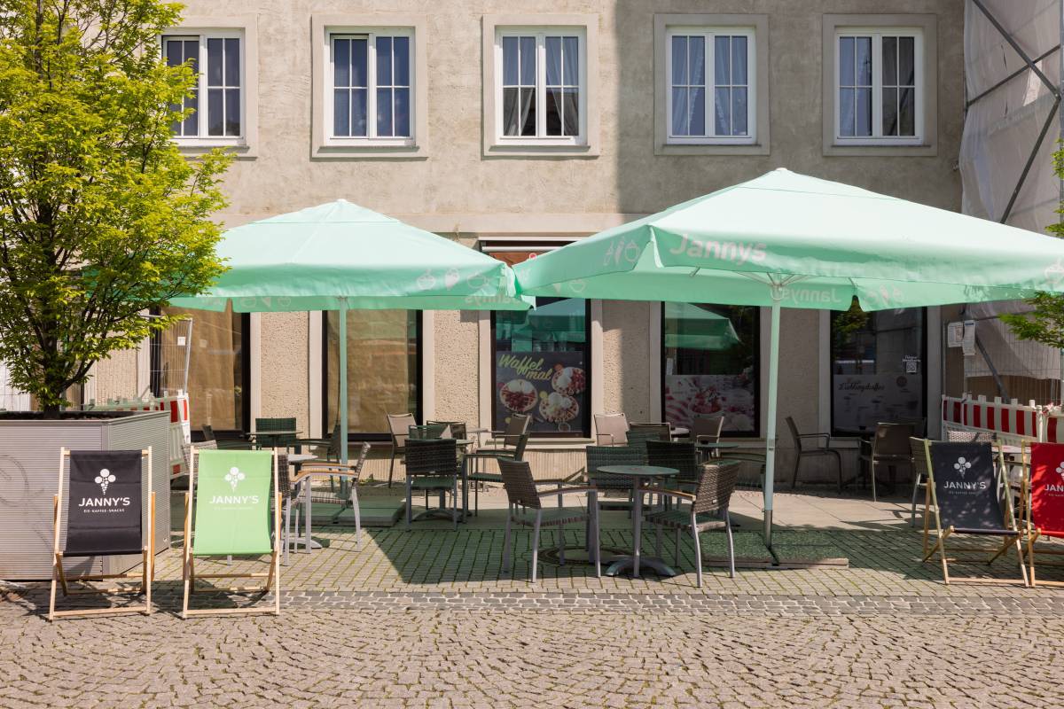 Beschirmter Außensitz- und Essbereich des Eiscafé Janny's Dessau mit Liegestühlen im Vordergrund und leichter Begrünung.