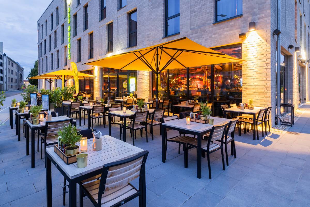 Beleuchteter Außensitz- und Essbereich des Restaurant Peter Pane Dessau in der Dämmerung mit gelben Sonnenschirmen und heller Sitzgarnitur aus Holz mit Tischdekoration