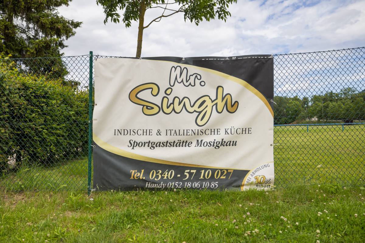 Begrünte Nahaufnahme vom Restaurantbanner am Zaun vor der Einfahrt mit großem Schriftzug Mr. Singh