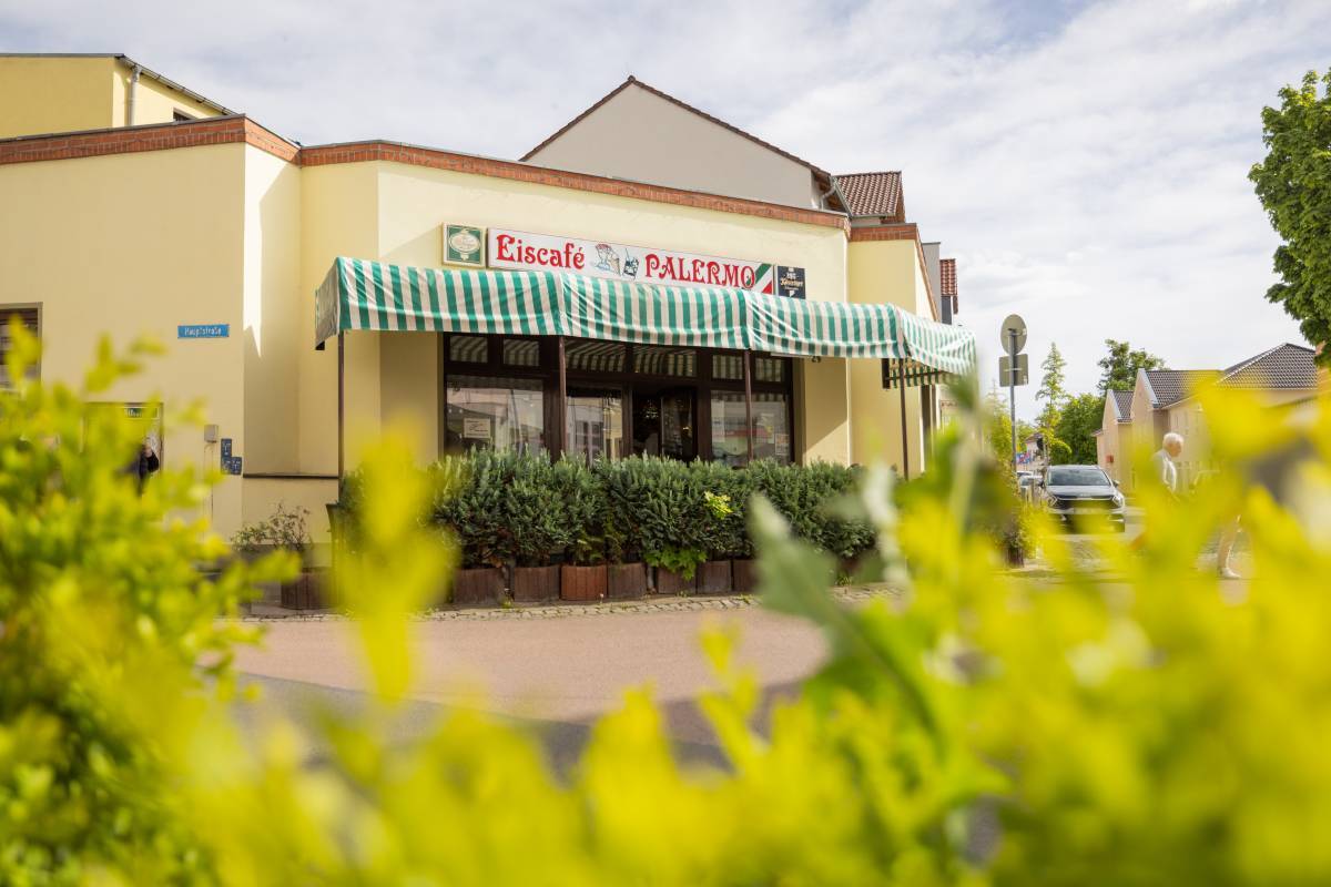 Begrünte Sicht auf den Eingangsbereich des Eiscafé Palermo Dessau mit grün-weiß gestreifter Markise und großer Fensterfront.