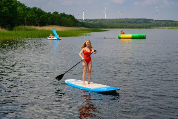 Eine junge Frau steht auf einem SUP, im Hintergrund sind ein Wassertrampolin und eine Tretboot zu sehen